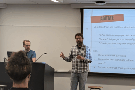 Two people at the front of a classroom, one behind a podium, the other gesturing while standing in front of a projected presentation with the word "AGITATE" in bold lettering on the screen. The back of another person facing them can be seen in the foreground.